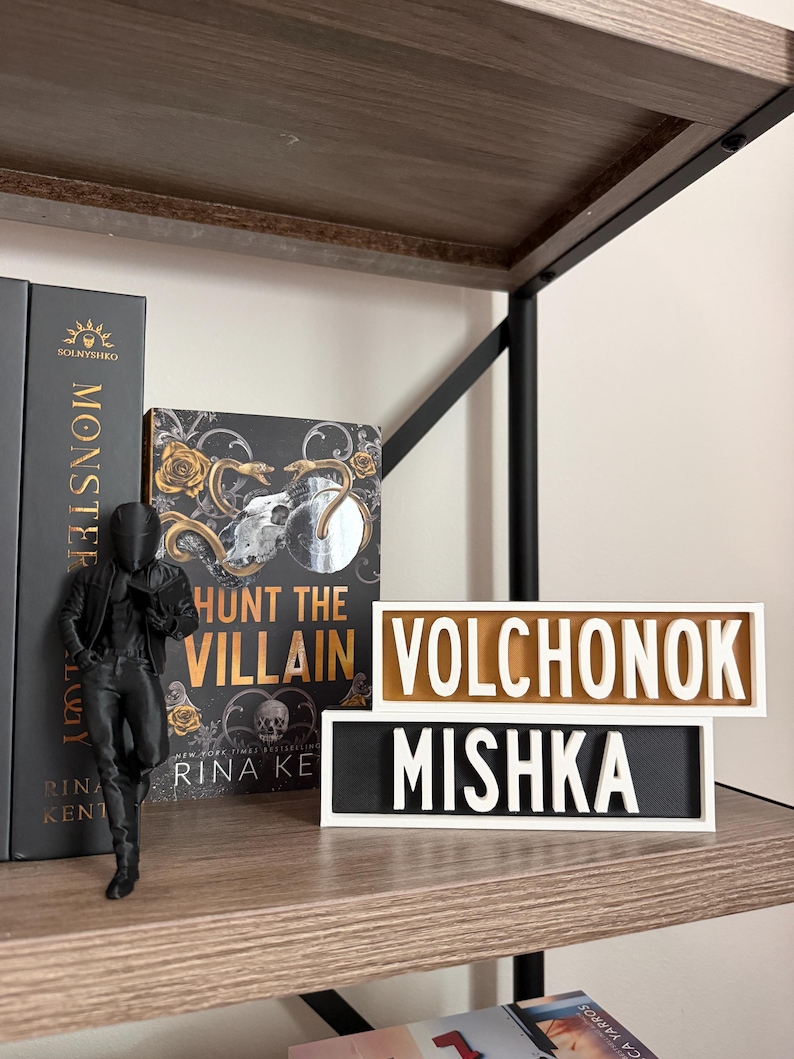 Puede incluir: Una estanter&iacute;a de madera con estructura met&aacute;lica negra contiene una figura, libros y dos letreros rectangulares. Los letreros dicen "VOLCHONOK" y "MISHKA" en letras blancas sobre un fondo negro y dorado. Una portada de libro dice "Hunt the Villain".