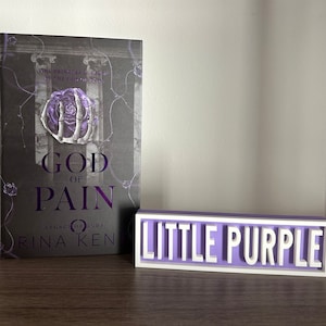 Może przedstawiać: Książka zatytułowana "God of Pain" z fioletowo-białą okładką, obok małego, prostokątnego znaku z napisem "Little Purple" białymi literami. Okładka książki zawiera wz&oacute;r r&oacute;ży i szkieletu dłoni.