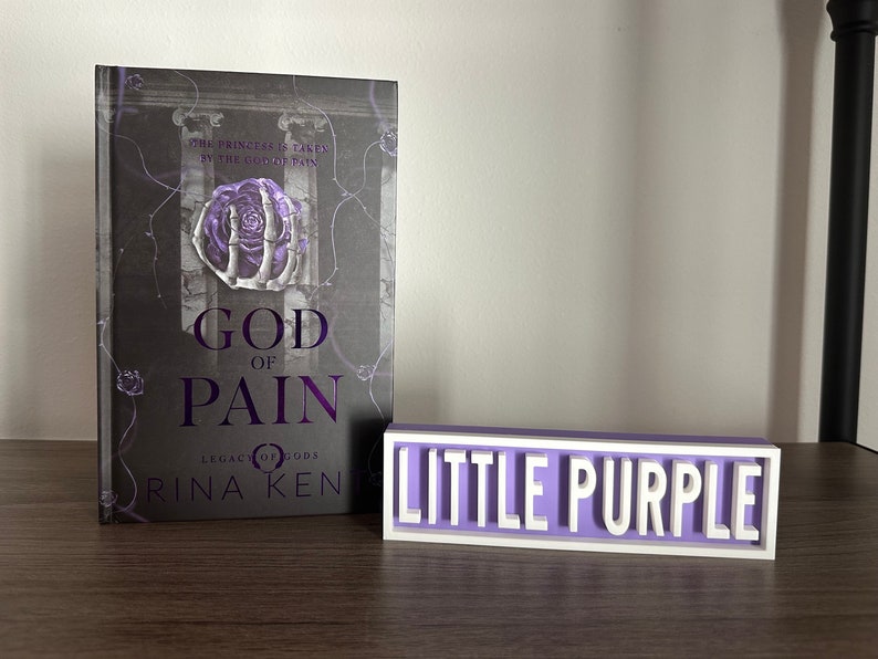 Może przedstawiać: Książka zatytułowana "God of Pain" z fioletowo-białą okładką, obok małego, prostokątnego znaku z napisem "Little Purple" białymi literami. Okładka książki zawiera wz&oacute;r r&oacute;ży i szkieletu dłoni.