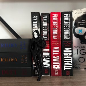 Puede incluir: Una figura negra de la muerte con rostro de calavera se encuentra junto a una fila de libros. Los libros tienen t&iacute;tulos como "Hideaway", "Kill Switch" y "Nightfall" de Penelope Douglas. Los libros est&aacute;n en una estanter&iacute;a de madera.