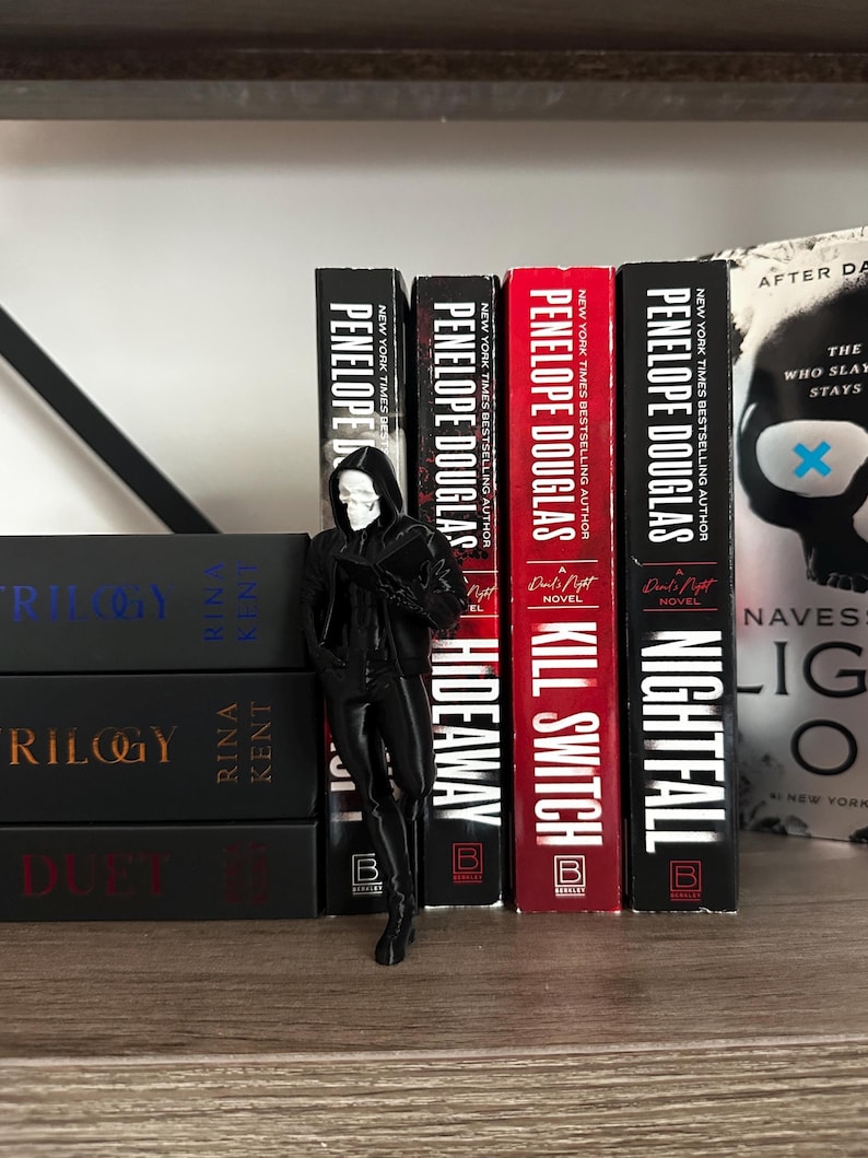 Puede incluir: Una figura negra de la muerte con rostro de calavera se encuentra junto a una fila de libros. Los libros tienen t&iacute;tulos como "Hideaway", "Kill Switch" y "Nightfall" de Penelope Douglas. Los libros est&aacute;n en una estanter&iacute;a de madera.