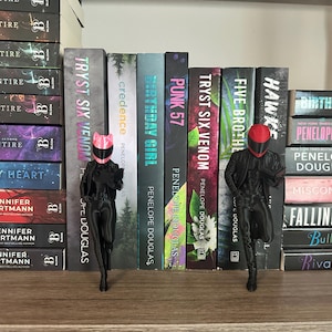 May include: A shelf displays a collection of books with titles like "Tryst Six Venom" and "Birthday Girl." Two black figurines, one with a pink cat-ear helmet and the other with a red helmet, stand in front of the books. The books are stacked on a wooden shelf.
