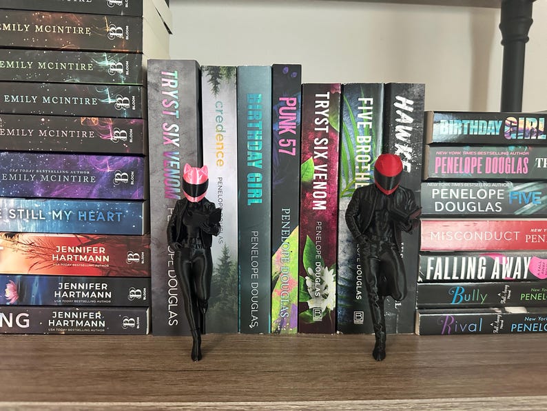 May include: A shelf displays a collection of books with titles like "Tryst Six Venom" and "Birthday Girl." Two black figurines, one with a pink cat-ear helmet and the other with a red helmet, stand in front of the books. The books are stacked on a wooden shelf.