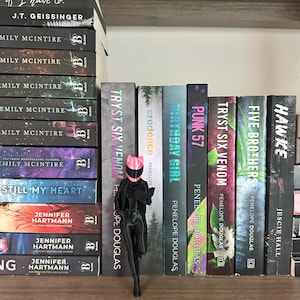May include: A stack of books with titles like "If I Have To" and "Still My Heart" sits on a wooden shelf. A black figurine wearing a pink helmet and holding a book stands in front of other books.