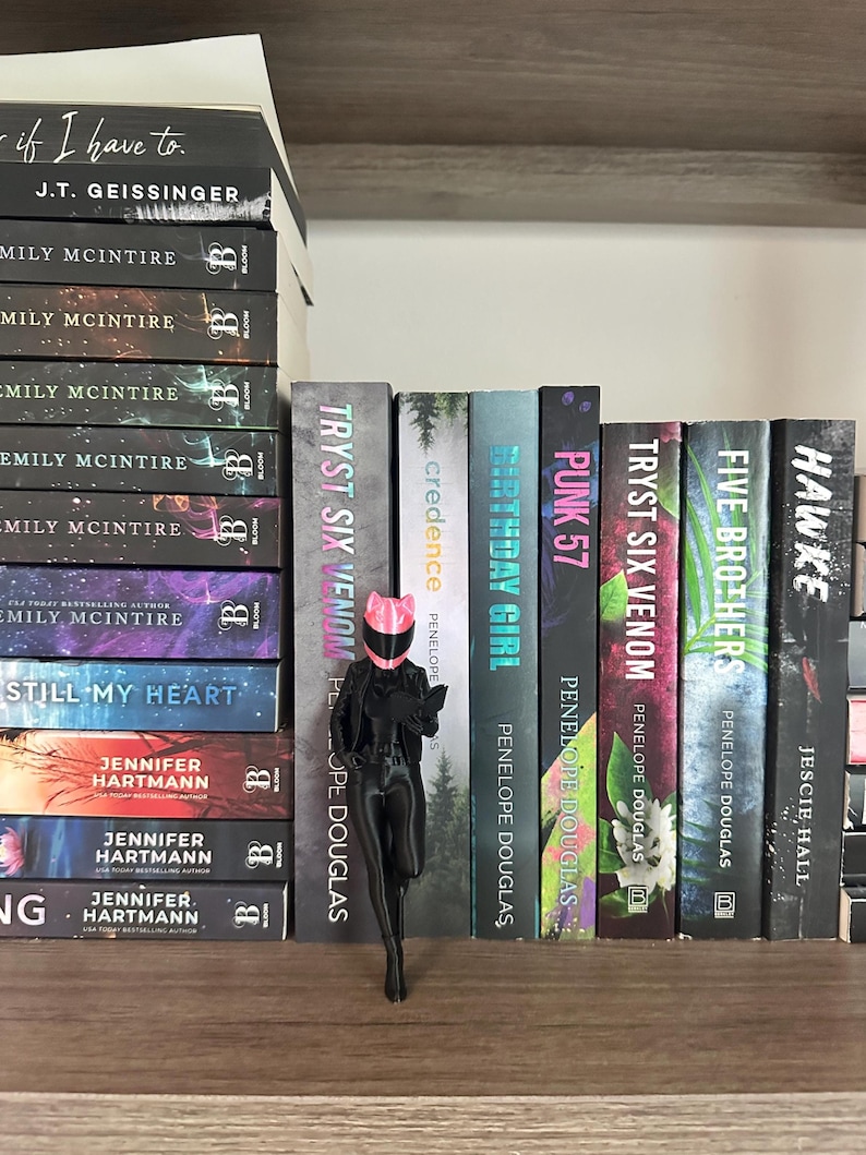 May include: A stack of books with titles like "If I Have To" and "Still My Heart" sits on a wooden shelf. A black figurine wearing a pink helmet and holding a book stands in front of other books.