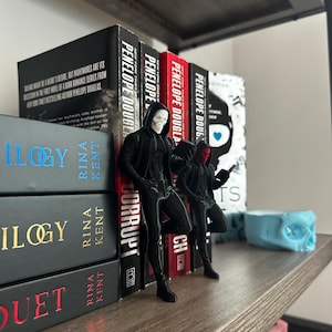 Puede incluir: Una estanter&iacute;a de madera con libros y figuras. Los libros son de Rina Kent y Penelope Douglas. Dos figuras negras con capuchas y m&aacute;scaras est&aacute;n cerca de los libros. Tambi&eacute;n se ve una calavera azul.