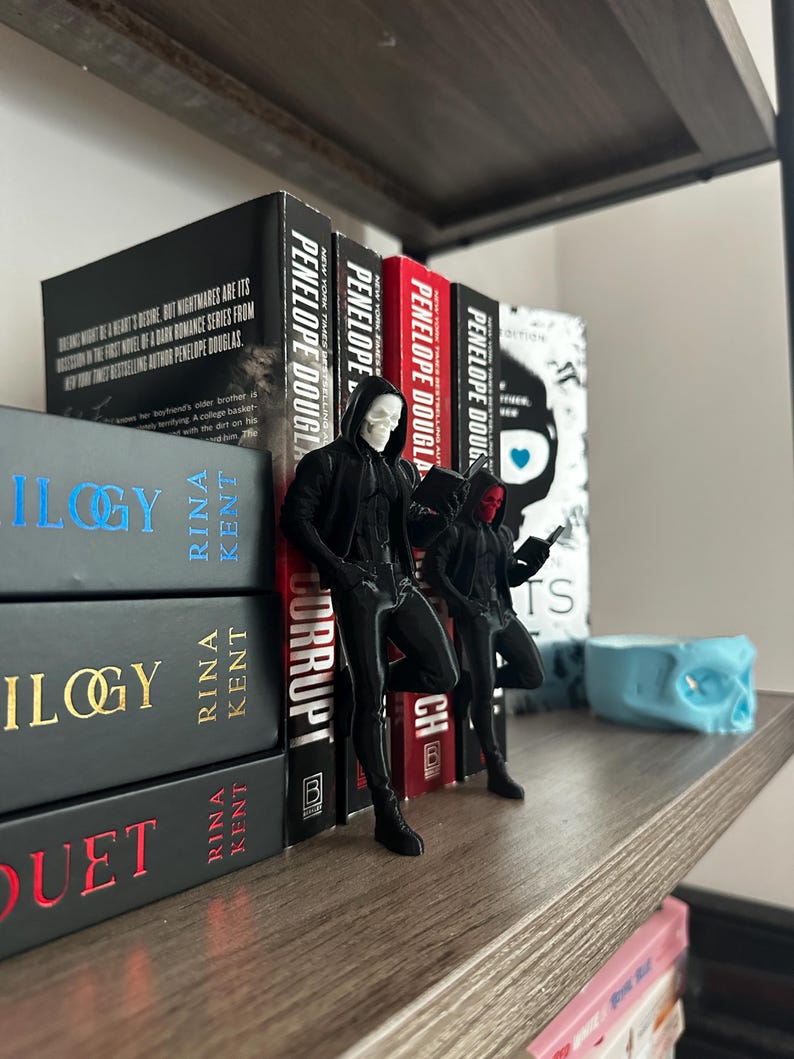 Puede incluir: Una estanter&iacute;a de madera con libros y figuras. Los libros son de Rina Kent y Penelope Douglas. Dos figuras negras con capuchas y m&aacute;scaras est&aacute;n cerca de los libros. Tambi&eacute;n se ve una calavera azul.