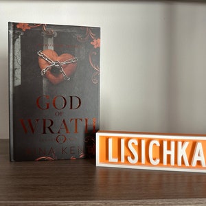 Może przedstawiać: Książka zatytułowana "God of Wrath" ze zdjęciem serca w łańcuchach na okładce oraz spersonalizowany znak z napisem "LISICHKA" białymi literami na pomarańczowym tle. Książka i znak znajdują się na drewnianej p&oacute;łce.