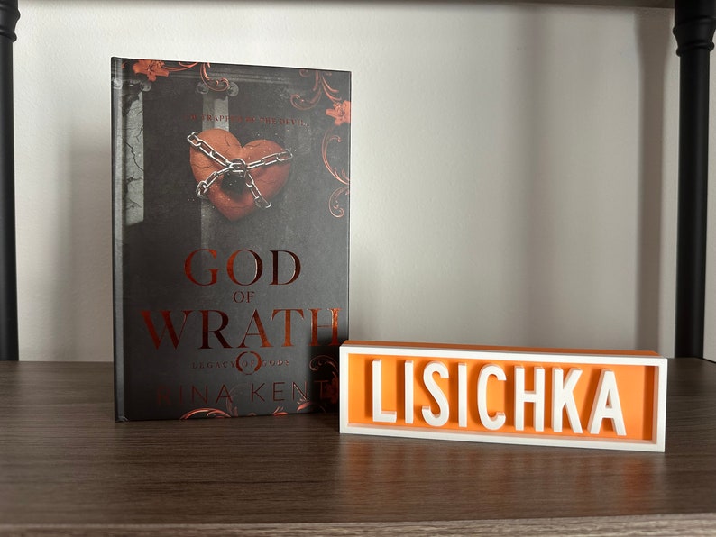 Może przedstawiać: Książka zatytułowana "God of Wrath" ze zdjęciem serca w łańcuchach na okładce oraz spersonalizowany znak z napisem "LISICHKA" białymi literami na pomarańczowym tle. Książka i znak znajdują się na drewnianej p&oacute;łce.