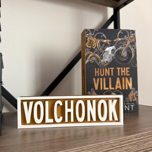 Puede incluir: Un letrero de madera con el nombre "VOLCHONOK" en letras blancas sobre un fondo marr&oacute;n, enmarcado en blanco. Un libro titulado "HUNT THE VILLAIN" con una portada dorada y negra est&aacute; detr&aacute;s del letrero, sobre una estanter&iacute;a de madera.
