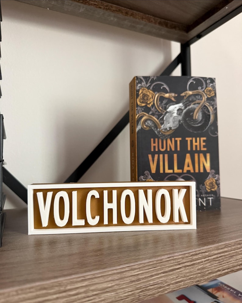 Puede incluir: Un letrero de madera con el nombre "VOLCHONOK" en letras blancas sobre un fondo marr&oacute;n, enmarcado en blanco. Un libro titulado "HUNT THE VILLAIN" con una portada dorada y negra est&aacute; detr&aacute;s del letrero, sobre una estanter&iacute;a de madera.
