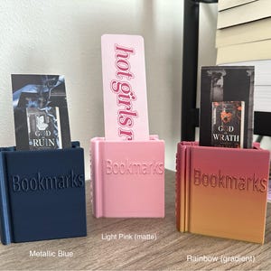 May include: Three bookmark holders in different colors: metallic blue, light pink, and rainbow gradient. Each holder has the word "Bookmarks" printed on the front.