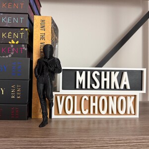 Puede incluir: Una figura negra de una persona con traje est&aacute; junto a dos placas de madera con las palabras "MISHKA VOLCHONOK" en letras blancas. Se ve una pila de libros con el nombre del autor "RINA KENT".