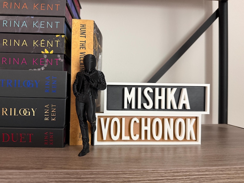 Puede incluir: Una figura negra de una persona con traje est&aacute; junto a dos placas de madera con las palabras "MISHKA VOLCHONOK" en letras blancas. Se ve una pila de libros con el nombre del autor "RINA KENT".