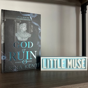 Może przedstawiać: Książka zatytułowana "God of Ruin" z ilustracją popiersia i tekstem "Legacy of Gods" i "Rina Kent". Obok znajduje się jasnoniebieski i biały znak z napisem "Little Muse".