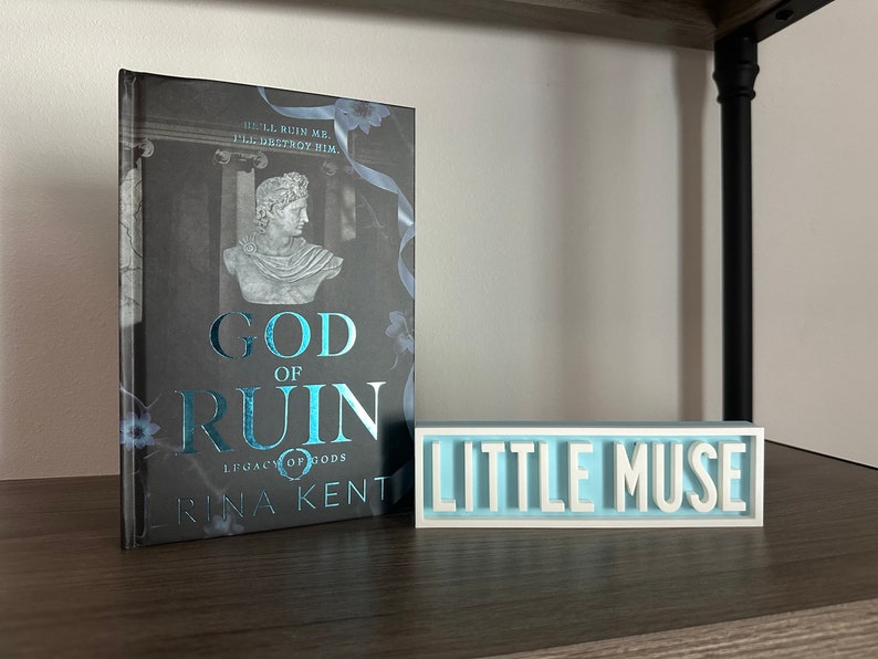 Może przedstawiać: Książka zatytułowana "God of Ruin" z ilustracją popiersia i tekstem "Legacy of Gods" i "Rina Kent". Obok znajduje się jasnoniebieski i biały znak z napisem "Little Muse".