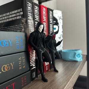 Puede incluir: Una estanter&iacute;a de madera con libros y figuras decorativas. Los libros son de Penelope Douglas y Rina Kent. Dos figuras negras con m&aacute;scaras de calavera est&aacute;n cerca de los libros. Tambi&eacute;n hay un recipiente azul claro con forma de calavera.