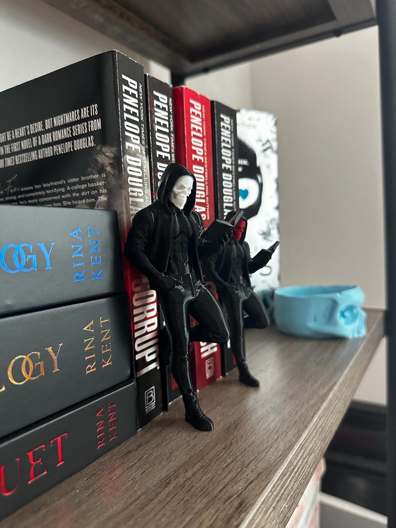 Puede incluir: Una estanter&iacute;a de madera con libros y figuras decorativas. Los libros son de Penelope Douglas y Rina Kent. Dos figuras negras con m&aacute;scaras de calavera est&aacute;n cerca de los libros. Tambi&eacute;n hay un recipiente azul claro con forma de calavera.