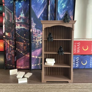 May include: A brown wooden bookshelf with three shelves. The top shelf has a black figurine on it. The middle shelf has a white book on it. The bottom shelf is empty. The bookshelf is sitting on a table with a brown surface. There are several books on the table behind the bookshelf.