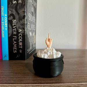 Op de afbeelding: Een zwarte ketel met een witte, schuimige substantie erin. Een hand met een opgestoken middelvinger steekt uit het schuim.