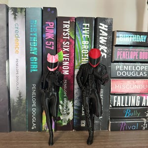 May include: Two black figurines of figures in motorcycle gear, one with a pink helmet and the other with a red helmet, stand in front of a bookshelf. The bookshelf contains several books with titles like "Birthday Girl" and "Punk 57".