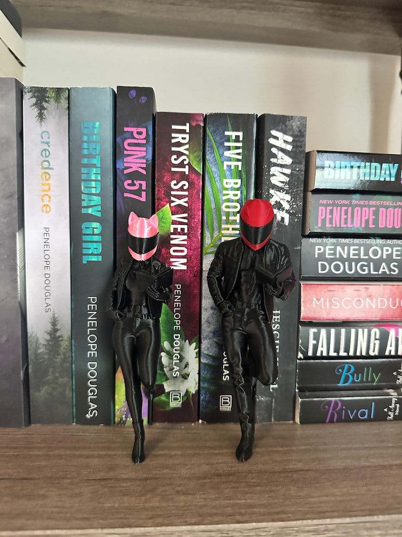 May include: Two black figurines of figures in motorcycle gear, one with a pink helmet and the other with a red helmet, stand in front of a bookshelf. The bookshelf contains several books with titles like "Birthday Girl" and "Punk 57".
