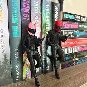 May include: Two black figurines of people wearing motorcycle helmets and jackets, reading books, are positioned in front of a shelf of books. The books have titles like "Birthday Girl" and "Punk 57" by Penelope Douglas.