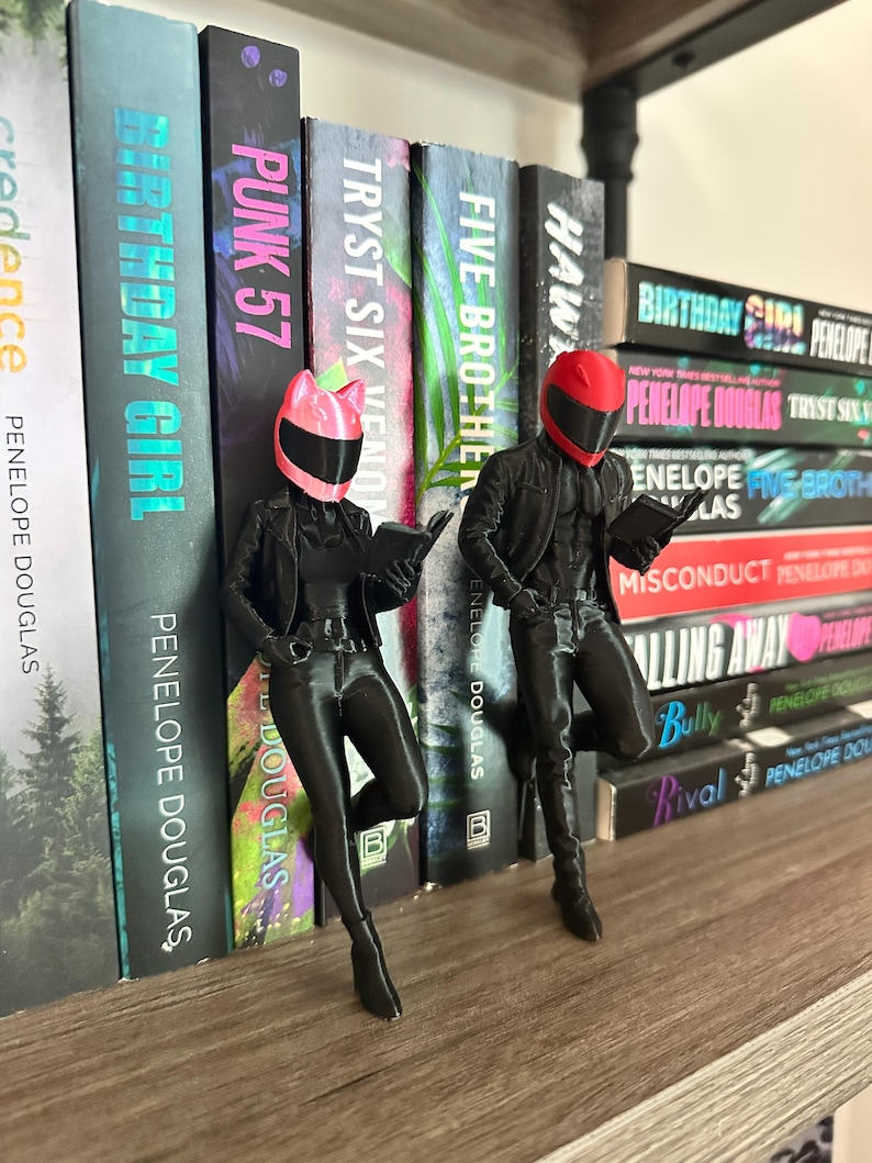 May include: Two black figurines of people wearing motorcycle helmets and jackets, reading books, are positioned in front of a shelf of books. The books have titles like "Birthday Girl" and "Punk 57" by Penelope Douglas.