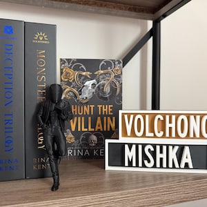Puede incluir: Una estanter&iacute;a con libros, incluyendo "Kingdom Duet" y "Monster". Una figura negra se apoya en los libros. Dos letreros de madera muestran las palabras "VOLCHONOK" y "MISHKA".