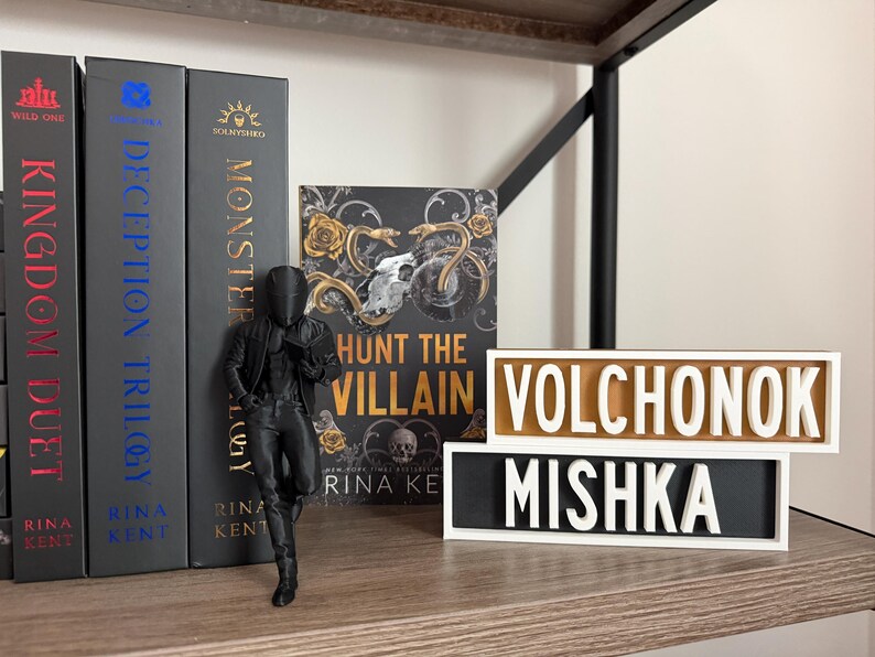 Puede incluir: Una estanter&iacute;a con libros, incluyendo "Kingdom Duet" y "Monster". Una figura negra se apoya en los libros. Dos letreros de madera muestran las palabras "VOLCHONOK" y "MISHKA".