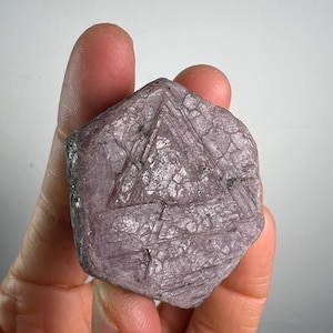 May include: A hexagonal, dusty purple raw crystal specimen featuring a prominent triangular geometric pattern etched into its surface. The stone has a matte, textured appearance with visible striations and crystalline formations across its face.