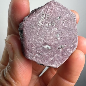 May include: A raw, hexagonal-shaped ruby crystal with a dusty pink and mauve hue. The surface displays a natural, textured finish with visible striations, geometric patterns, and small, embedded dark mineral inclusions across its flat face.