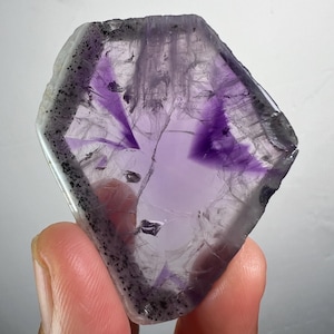 May include: A translucent, irregularly shaped amethyst crystal with a deep purple centre and a clear, crystalline structure. The edges are lined with a dark, speckled border. The crystal is held in a person's hand.