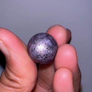 May include: A spherical, polished gemstone with a translucent, grayish-purple hue. The surface displays a distinct six-rayed star pattern radiating from the center, accompanied by scattered dark, irregular inclusions throughout the stone's body.