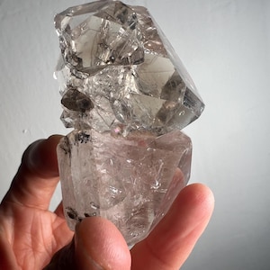 Op de afbeelding: Een heldere kwartskristalcluster met een rokerige tint. De kristal wordt in een hand gehouden en toont zijn natuurlijke, veelzijdige structuur en doorschijnende kwaliteit. Het oppervlak van de kristal reflecteert licht.