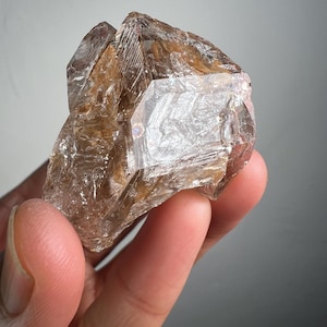 May include: A raw, natural quartz crystal with a translucent, light brown hue. The crystal has multiple facets and a rough, textured surface. The light catches the edges, creating a shimmering effect. The crystal is held in a hand.