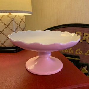 Puede incluir: Un soporte para tartas de cerámica rosa y blanco con un borde festoneado. El soporte tiene una base de pedestal.