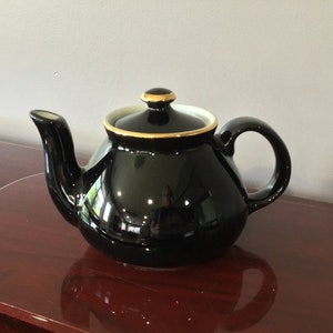 Puede incluir: Una tetera negra con un borde dorado alrededor de la tapa y un interior blanco. La tetera tiene un cuerpo redondeado y un pico curvo.