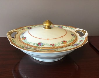 May include: A white porcelain serving bowl with a gold rim and floral pattern. The bowl has two handles and a lid with a gold knob.