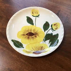 May include: A white ceramic plate with a yellow and brown floral design. The plate features a large yellow flower with brown center and smaller yellow flowers with brown centers. The flowers are surrounded by green leaves.