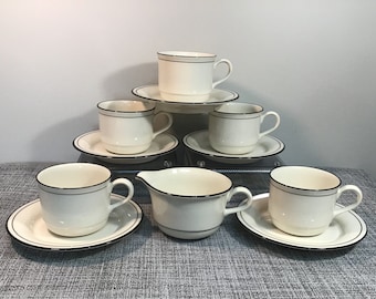 May include: Set of six white ceramic teacups and saucers with a black trim. The set also includes a matching creamer.