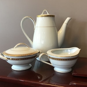 Puede incluir: Un juego de cafetera, jarra de crema y azucarero de porcelana blanca con ribetes dorados. El juego presenta un delicado patrón floral.