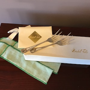 Puede incluir: Un juego de dos tenedores de plata en una caja blanca con una bolsa de tela verde. La caja tiene el texto "Marshall Field's" impreso en ella.