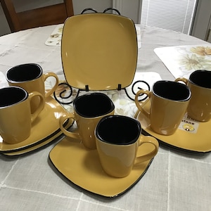 May include: Set of six yellow ceramic mugs with black rims and matching square plates. The mugs and plates have a modern design.