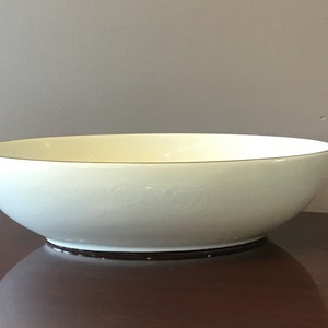 Pode incluir: Uma tigela grande de cerâmica branca com forma oval ligeiramente arredondada. A tigela tem uma fina borda dourada e um design em relevo subtil.