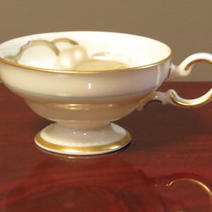 Può includere: Una tazza da tè bianca con un bordo dorato e una fascia blu intorno al centro. La tazza ha un manico delicato e si trova su un piccolo piedistallo.