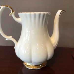May include: White porcelain teapot with a gold trim around the rim and base. The teapot has a fluted design and a curved handle.