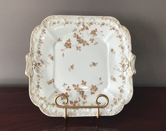 May include: A white porcelain serving platter with a gold rim and a floral pattern in brown and gold. The platter has a square shape with rounded corners and a raised edge.