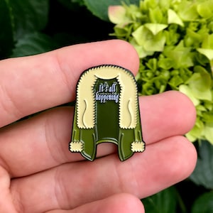 May include: Green and cream enamel pin shaped like a cardigan with the text "It's all happening" on the front.