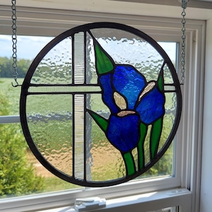 May include: Round stained glass window art featuring a vibrant blue iris flower with green leaves. The flower is set against a clear, textured background with a black metal frame. The window hangs from a chain.